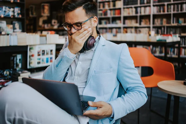 Orta yaşlı yakışıklı adam modern kitapçıda oturuyor, kulaklıkla müzik dinlerken dizüstü bilgisayarını ya da bilgisayarını okuyor ve kullanıyor..