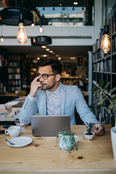 Genç yakışıklı adam modern kitapçıda oturuyor, dizüstü bilgisayarını veya bilgisayarını okuyor ve kullanıyor..