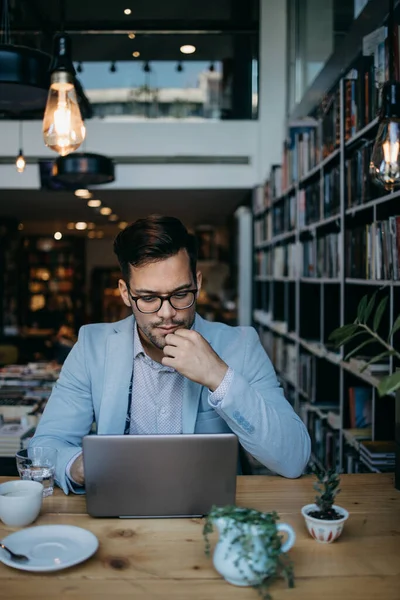Genç yakışıklı adam modern kitapçıda oturuyor, dizüstü bilgisayarını veya bilgisayarını okuyor ve kullanıyor..