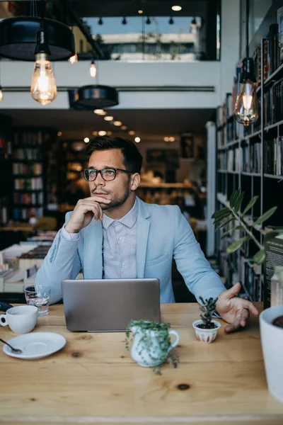 Genç yakışıklı adam modern kitapçıda oturuyor, dizüstü bilgisayarını veya bilgisayarını okuyor ve kullanıyor..