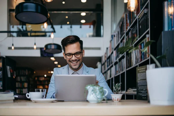 Genç yakışıklı adam modern kitapçıda oturuyor, dizüstü bilgisayarını veya bilgisayarını okuyor ve kullanıyor..