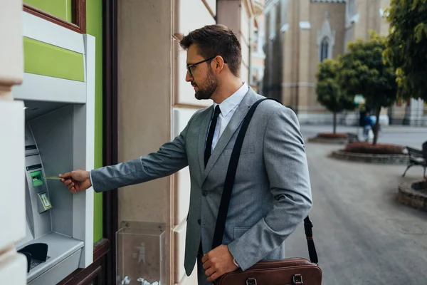 Handsome middle age businessman with eyeglasses standing on city street and using ATM machine to withdraw money from credit or debit card. 