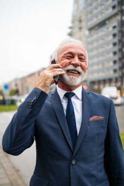 Zarif ve pozitif kıdemli işadamı caddede duruyor ve akıllı telefonunu kullanıyor. Düşük açı görünümü. 