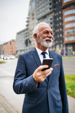 Zarif ve pozitif kıdemli işadamı caddede duruyor ve akıllı telefonunu kullanıyor. Düşük açı görünümü. 