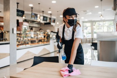 Beautiful woman working bakery or fast food restaurant. She is cleaning and disinfecting tables against Coronavirus pandemic disease. She is wearing protective face masks and gloves.