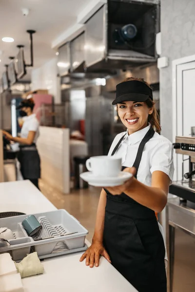 Beautiful and happy young female worker working in a bakery or fast food restaurant and using coffee machine. Positive people in small business concept.