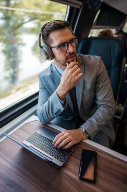 Hızlı trenle seyahat ederken dizüstü bilgisayarını kullanan orta yaşlı yakışıklı bir iş adamı. Düşünceli bir şekilde yana bakıyor. Modern ve hızlı seyahat kavramı. Yüksek açı görünümü.