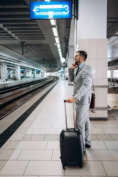 Handsome middle age businessman with suitcase standing in station platform and waiting for high-speed train or metro. Modern and fast travel concept.