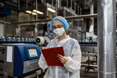 Tıbbi malzeme araştırma ve üretim fabrikasında çalışan ve sevkiyattan önce damıtılmış su kutularını kontrol eden koruyucu yüz maskeli bir kadın işçi. Denetim kalite denetimi. 