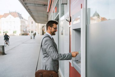 Handsome middle age businessman with eyeglasses standing on city street and using ATM machine to withdraw money from credit or debit card.