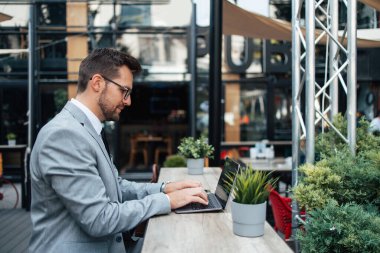 Orta yaşlı işadamı kafeteryada oturuyor ve iki toplantı arasında kahve molası veriyor. Dizüstü bilgisayar kullanıyor ve pozitif ve kendinden emin..