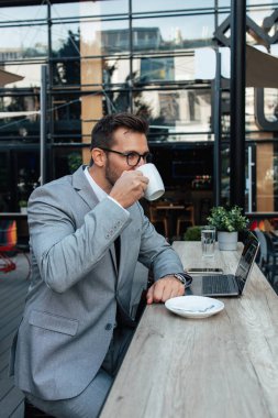 Orta yaşlı işadamı kafeteryada oturuyor ve iki toplantı arasında kahve molası veriyor. Dizüstü bilgisayar kullanıyor ve pozitif ve kendinden emin.. 
