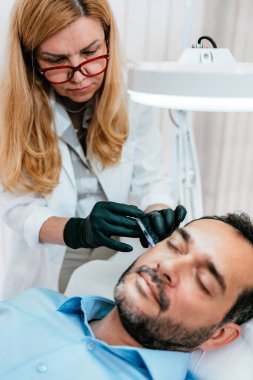 Yakışıklı orta yaşlı sakallı adam güzellik kliniğinde gençleştirici yüz enjeksiyonu yaptırıyor. Güzellik uzmanı erkek kırışıklıklarını botulinyum toksin iğneleriyle ya da hyaluronik asit doldurucularıyla dolduruyor..