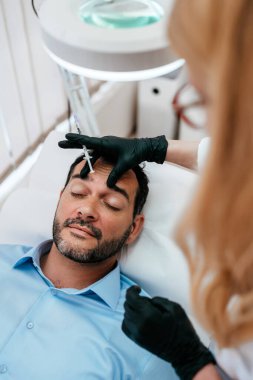 Yakışıklı orta yaşlı sakallı adam güzellik kliniğinde gençleştirici yüz enjeksiyonu yaptırıyor. Güzellik uzmanı erkek kırışıklıklarını botulinyum toksin iğneleriyle ya da hyaluronik asit doldurucularıyla dolduruyor..
