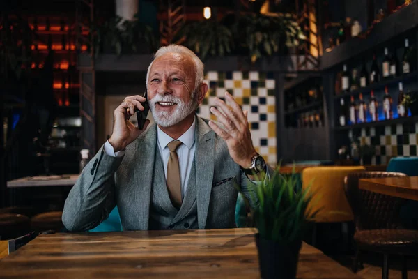 Happy businessman sitting in restaurant and waiting for lunch. He is using smart phone and talking with someone. Business seniors lifestyle concept.