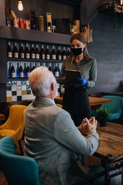 Waitress serves and takes the order from the senior businessman at the restaurant. She wears a protective mask as part of security measures against the Coronavirus pandemic.