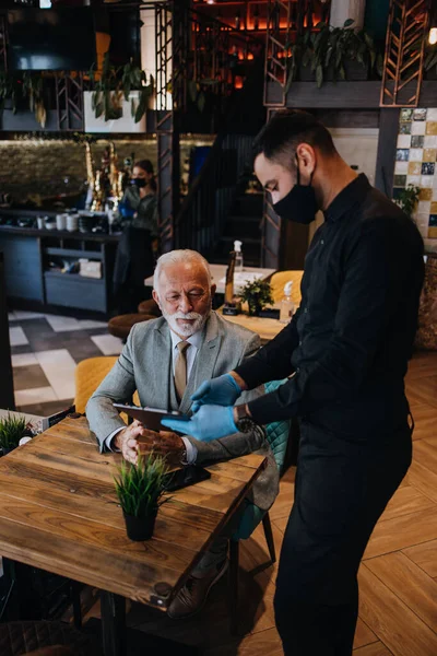 The waiter taking order for a delicious meal to the senior businessman at the restaurant. He wears a protective mask as part of security measures against the Coronavirus pandemic.