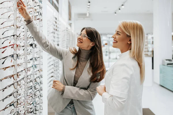 Modern optik mağazada gözlük çerçevesi seçen güzel ve modaya uygun bir kadın. Kadın satıcı uzmanı doğru karar vermesine yardımcı oluyor..