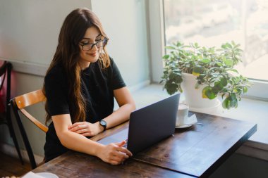 Gözlüklü genç kadın kafede oturuyor, kahve içiyor ve dizüstü bilgisayarla çalışıyor.