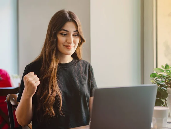 Online galibiyeti kutlayan heyecanlı genç bayan kafeye dizüstü bilgisayarla bakıyor..