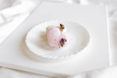 Pembe kalp şeklinde bir pasta beyaz bir tabakta, arka planda yatar. Beyaz bir tuval üzerinde hafif badem kurabiyeleri. Sevgililer Günü arkaplanı