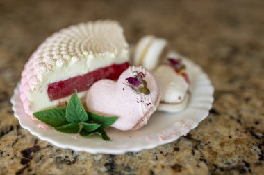 Beyaz-pembe bir kek parçası beyaz bir tabakta, açık bir arka planda yatıyor. Pembe kalp şeklinde bir pasta beyaz bir tabakta, arka planda yatar. Beyaz bir tuval üzerinde hafif badem kurabiyeleri. Lezzetli hamur işleri