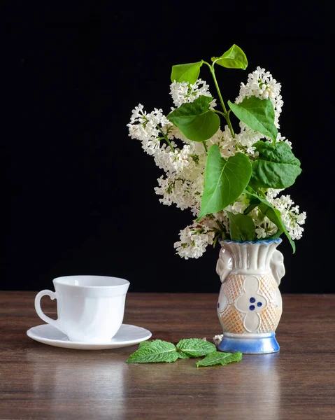 Masanın üzerinde vazoda beyaz bir leylak var. Yanında beyaz bir kupa var..