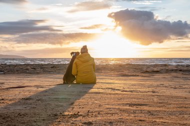 Bir Alman boksör köpeği gün batımında sahil kenarında oturuyor. Yanında sarışın takım arkadaşı İsveçli kız var..