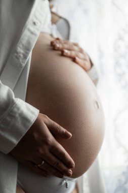 Güzel bir hamile göbeğine yakın çekim. Genç hamile bir kadın karnına dokunuyor, beyaz arka planda soyutlanmış. Hamile kadın, elleri sırtında karnını tutan hamile kadın..