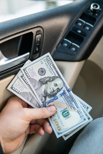 Ruce mladého muže držícího nové dolarové bankovky na pozadí nových aut. Koncept nákupu a prodeje automobilů — Stock fotografie