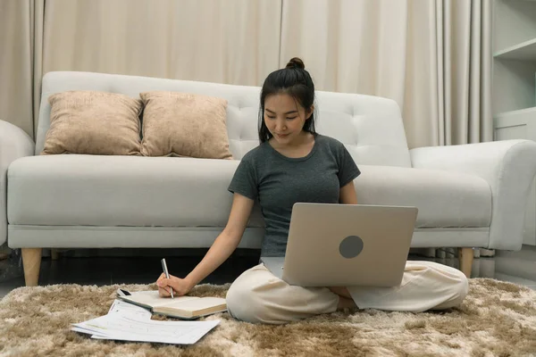 Azjatka Siedzi Dywanie Salonie Pracuje Komputerze Zapisując Notatki Notatniku — Zdjęcie stockowe