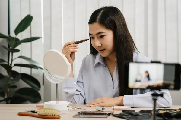 若いアジアの女性は 彼女のビデオカメラとテーブルの上の化粧品スキンケア製品についてVloggingと彼女のオンラインブログチャンネルのための製品の使用とレビューを実証します — ストック写真