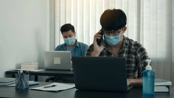 Asians sit in their office and use the phone to talk to clients while wearing masks in their offices during COVID-19.