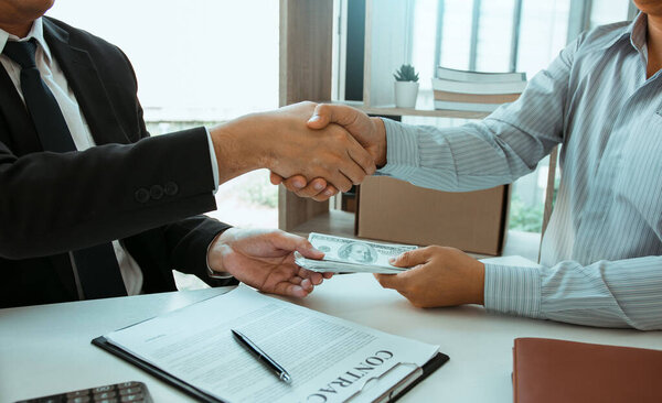 Два корпоративных бизнесмена пожимают друг другу руки, в то время как один человек кладет деньги на документ в офисе с коррупционной концепцией.