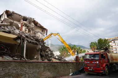 Alanya, Türkiye - 11 / 29 / 2022: Sarı bir kazıcı Türkiye 'nin Alanya kentindeki eski bir evi yıkmak için çalışıyor. Elinde su dolu bir varil olan bir adam, molozları tozsuz tutmak için üzerine su döküyor..
