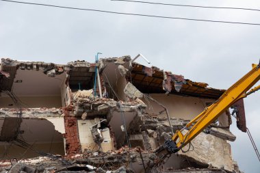 Türkiye 'nin Alanya kentindeki eski bir evi yıkmak için sarı bir kazıcı çalışıyor. Elinde su dolu bir varil olan bir adam, molozları tozsuz tutmak için üzerine su döküyor..