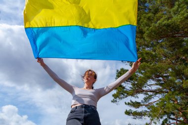 Ukrayna 'nın bayrağını taşıyan genç bir kadın, dağlara ve Ukrayna doğasına ve dramatik bulutlu gökyüzüne bakan güzel manzaranın arka planına karşı. Ukrayna Bayrağıyla Vatansever Kadın.