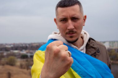 Ukrayna bayrağına sahip bir adam Ukrayna şehrinin arka planında ve gölünde yumruğunu gösteriyor. Ukrayna bayrağıyla Ukraynalı bir vatanseverin portresi.