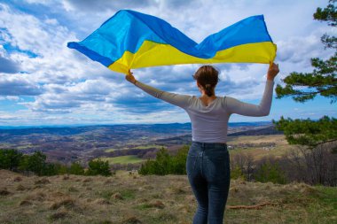 Ukrayna 'nın bayrağını taşıyan genç bir kadın, dağlara ve Ukrayna doğasına ve dramatik bulutlu gökyüzüne bakan güzel manzaranın arka planına karşı. Ukrayna Bayrağıyla Vatansever Kadın.