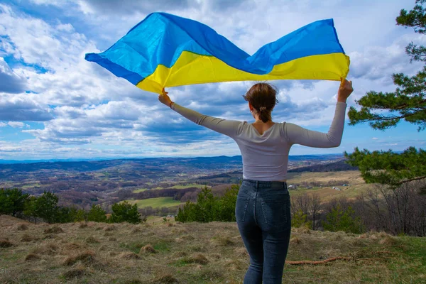 Ukrayna 'nın bayrağını taşıyan genç bir kadın, dağlara ve Ukrayna doğasına ve dramatik bulutlu gökyüzüne bakan güzel manzaranın arka planına karşı. Ukrayna Bayrağıyla Vatansever Kadın.