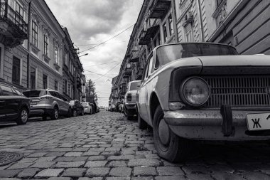 Ukrayna, Chernivtsi, 03.27.2022: Chernivtsi şehrinde park etmiş arabalı sokak. Eski araba Moskvich ön planda. Kenarları kaldırımlı şehir caddesi ve eski binalar. Siyah ve beyaz resim.