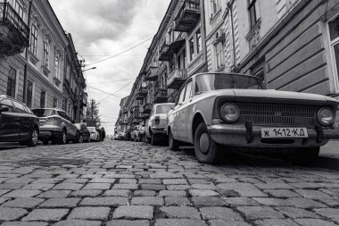 Ukrayna, Chernivtsi, 03.27.2022: Chernivtsi şehrinde park etmiş arabalı sokak. Eski araba Moskvich ön planda. Kenarları kaldırımlı şehir caddesi ve eski binalar. Siyah ve beyaz resim.
