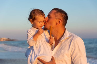 Küçük bir kızı olan genç bir baba gün batımında deniz kıyısında yürüyor. Beyaz elbiseli bir baba ve iki yaşındaki bir kız deniz dalgalarının arka planında sörf çizgisi boyunca yürüyorlar. Aile değerleri hakkında fotoğraf, Babalar Günü..