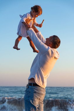 Küçük bir kızı olan genç bir baba gün batımında deniz kıyısında yürüyor. Beyaz elbiseli bir baba ve iki yaşındaki bir kız deniz dalgalarının arka planında sörf çizgisi boyunca yürüyorlar. Aile değerleri hakkında fotoğraf, Babalar Günü..