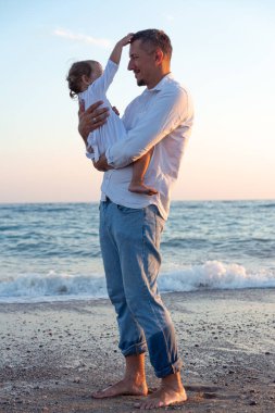 Küçük bir kızı olan genç bir baba gün batımında deniz kıyısında yürüyor. Beyaz elbiseli bir baba ve iki yaşındaki bir kız deniz dalgalarının arka planında sörf çizgisi boyunca yürüyorlar. Aile değerleri hakkında fotoğraf, Babalar Günü..