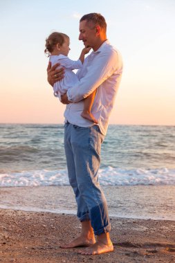 Küçük bir kızı olan genç bir baba gün batımında deniz kıyısında yürüyor. Beyaz elbiseli bir baba ve iki yaşındaki bir kız deniz dalgalarının arka planında sörf çizgisi boyunca yürüyorlar. Aile değerleri hakkında fotoğraf, Babalar Günü..