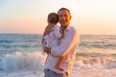 Küçük bir kızı olan genç bir baba gün batımında deniz kıyısında yürüyor. Beyaz elbiseli bir baba ve iki yaşındaki bir kız deniz dalgalarının arka planında sörf çizgisi boyunca yürüyorlar. Aile değerleri hakkında fotoğraf, Babalar Günü..