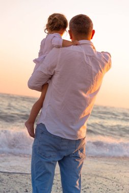 Küçük bir kızı olan genç bir baba gün batımında deniz kıyısında yürüyor. Beyaz elbiseli bir baba ve iki yaşındaki bir kız deniz dalgalarının arka planında sörf çizgisi boyunca yürüyorlar. Aile değerleri hakkında fotoğraf, Babalar Günü..