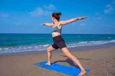 A beautiful girl is doing yoga on the beach near the sea. A girl in tracksuit does yoga exercises on the sand, and against the backdrop of the sea surf and a beautiful sky.