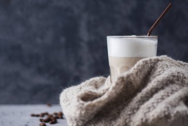 Bir bardak kahve kapuçinosunun yakın çekimi. Masanın üzerinde tarçın ve örgü bir eşarp var..